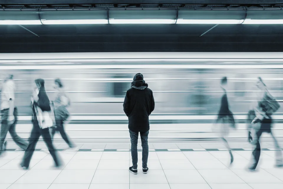 Monotone photo of man waiting on subway platform with blurry train passing by and other passengers walking by